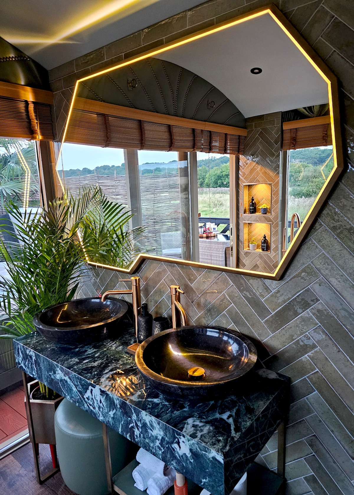A modern bathroom features a striking hexagonal mirror illuminated with soft backlighting above a double sink. The dark marble basin contrasts with the textured wall tiles. Large windows provide natural light and views of the surrounding landscape, complemented by touches of greenery.