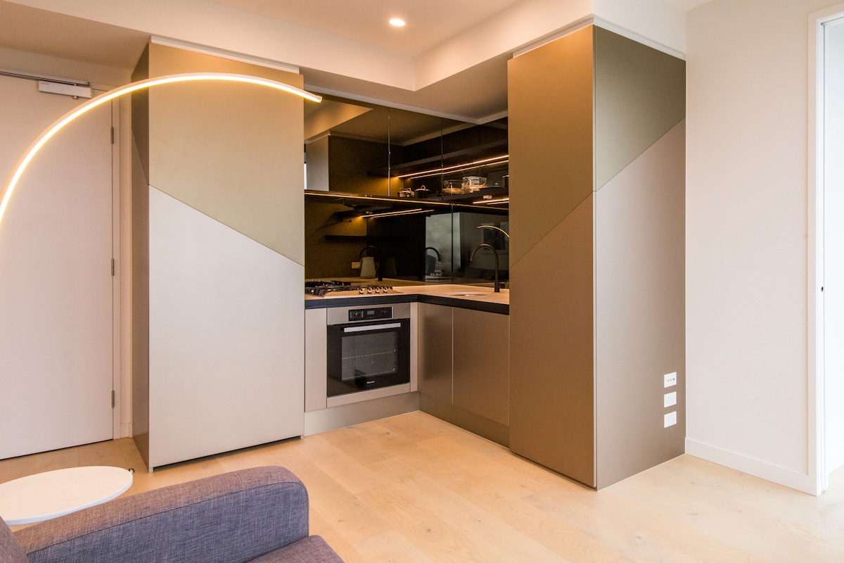 A modern kitchen is showcased with stylish, geometric cabinetry and sleek countertops. An integrated oven and stovetop are visible, along with minimalist shelving. Soft lighting enhances the contemporary design, while the wooden floor adds warmth to the overall aesthetic.