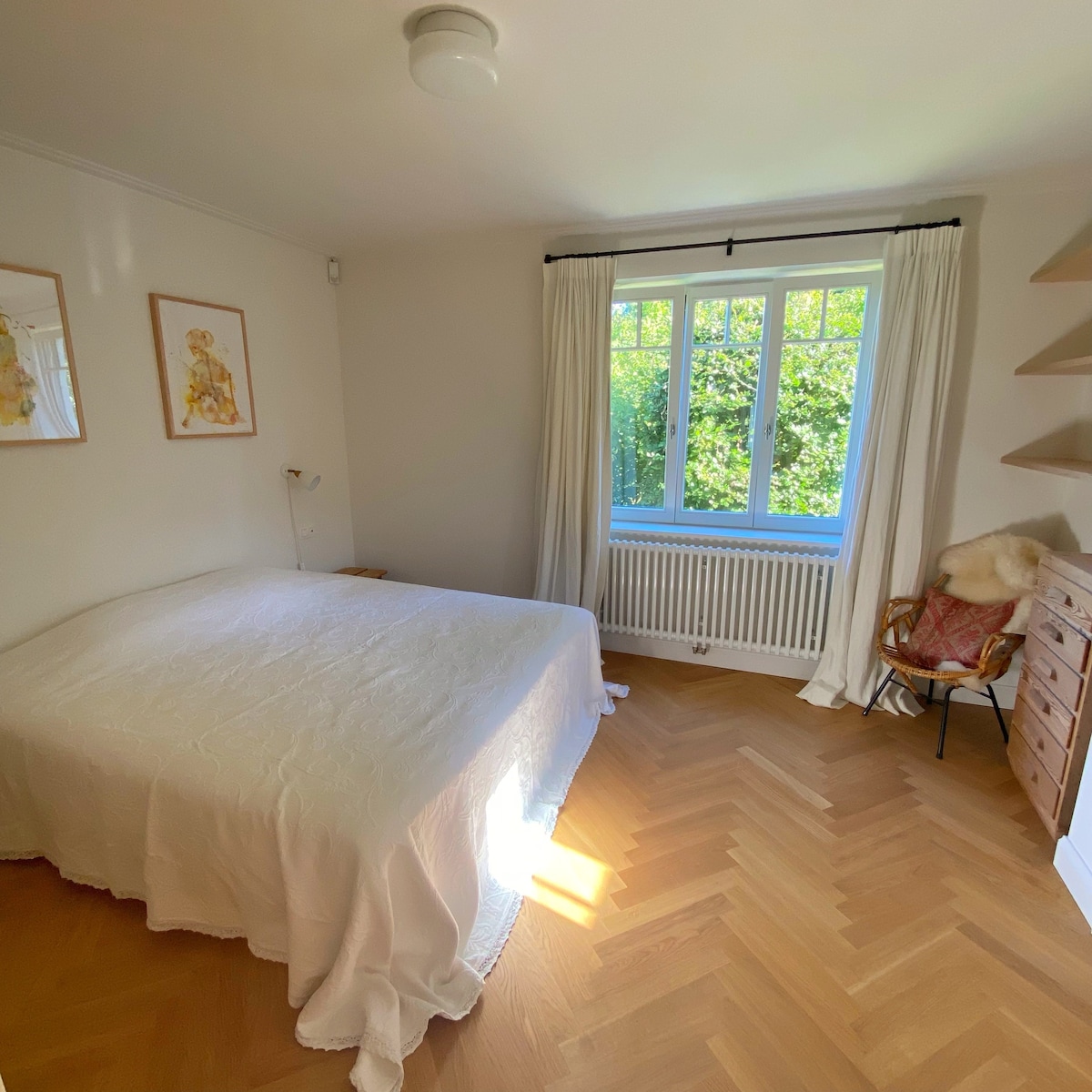 A bright bedroom features a spacious bed covered with a white quilt. Natural light illuminates the room through a large window, which offers views of greenery outside. Simple artwork decorates the walls, and wooden shelving adds practical touch.