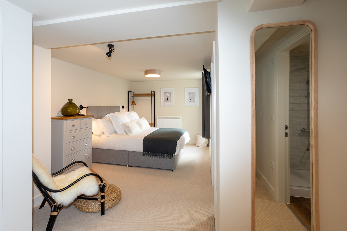 A spacious bedroom features a king-size bed with plush bedding, complemented by a patterned throw. A stylish cabinet stands beside the bed, with framed artwork adorning the walls. A doorway leads to a sleek shower room, partially visible in the background.