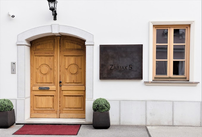The entrance features double wooden doors crafted with circular engravings, flanked by potted plants. A sign displaying 'Zabjak 5' is mounted on the wall beside a window with multiple panes, allowing natural light to illuminate the area.