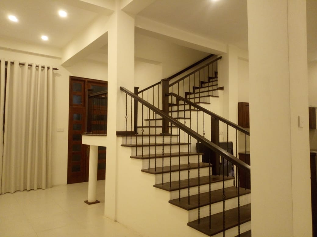 A spacious interior is showcased, featuring a staircase with dark wooden steps and a white railing. Soft lighting illuminates the area, highlighting the high ceilings and pale walls. A doorway with wooden paneling and curtains can also be seen in the background.