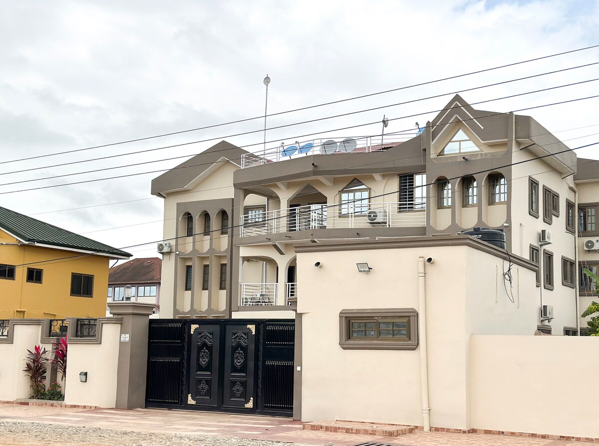 The exterior of a modern apartment building is presented, featuring a combination of light and dark tones. The entrance is marked by a stylish black gate, while balconies adorn the upper levels. A rooftop area with lounge chairs is visible, inviting relaxation.