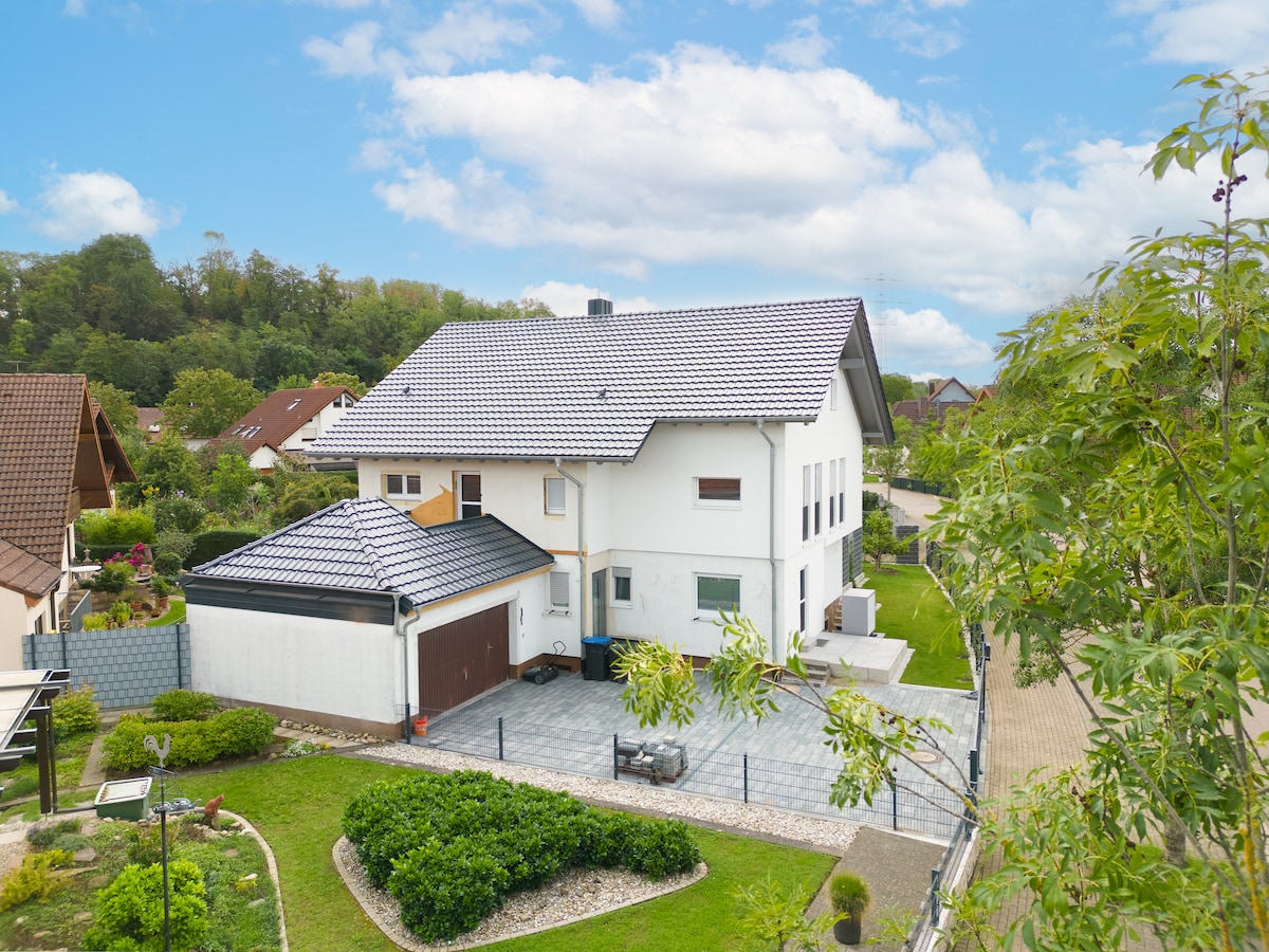 The exterior of the property is shown, highlighting a two-story house with a tiled roof. A well-maintained garden surrounds the building, featuring neatly trimmed shrubs and a paved area. The backdrop includes trees and other residential structures, contributing to a serene neighborhood atmosphere.