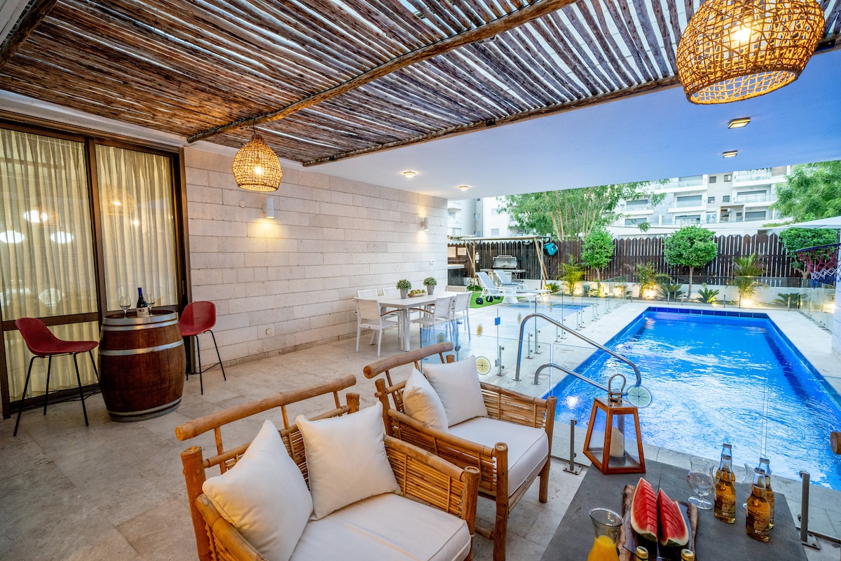 A shaded outdoor area features comfortable seating with white cushions and a wooden table. A private pool is visible, surrounded by lush greenery. The ambiance is enhanced by decorative light fixtures, while a dining table is set for outdoor meals nearby.