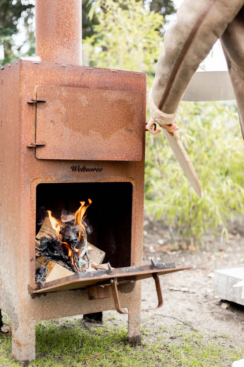 A rustic wood-burning stove is shown in operation, with flames emerging from the open door. The body of the stove is constructed from aged metal, showcasing a warm, textured appearance. A hand is seen adjusting the wood being added for continued burning.