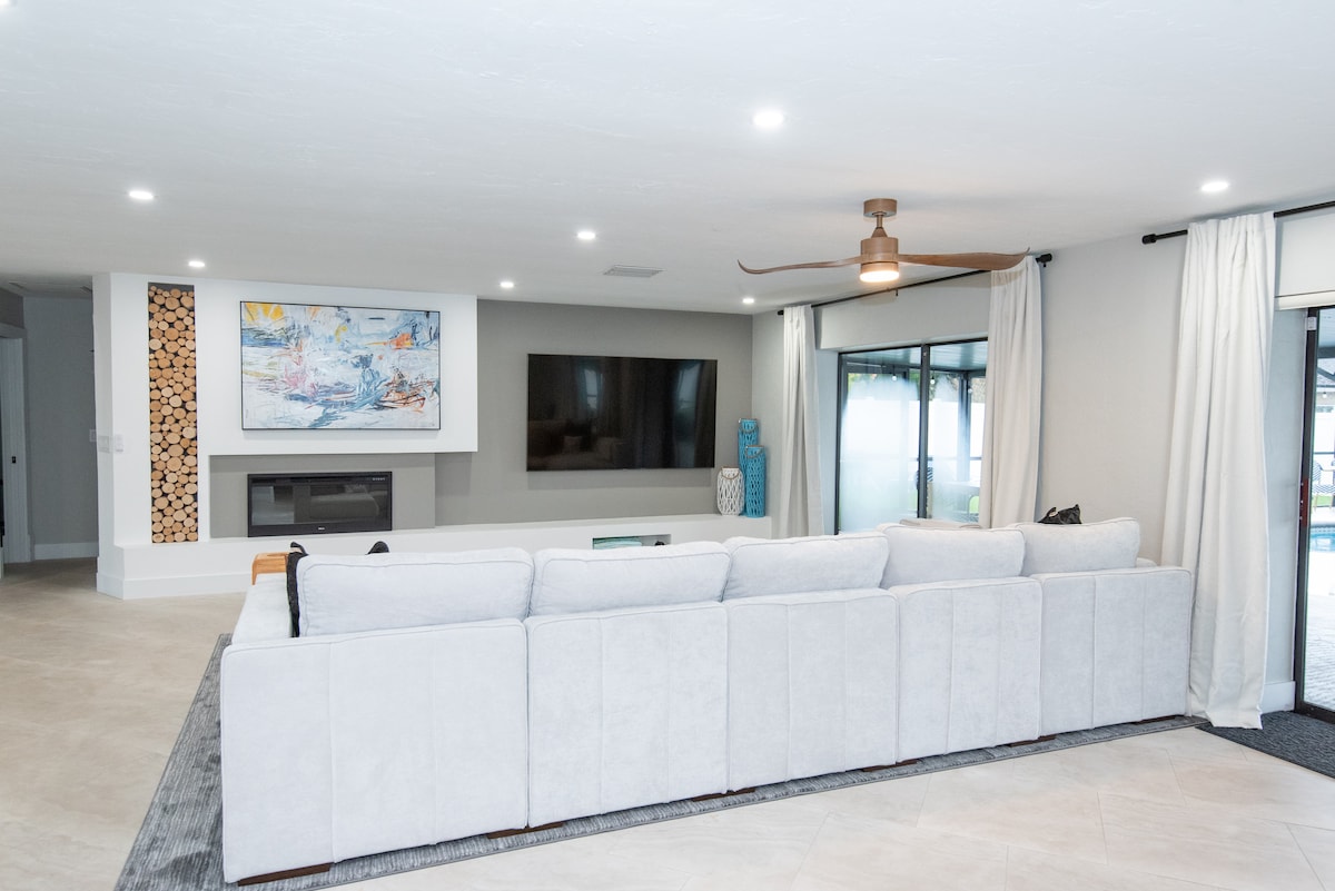 A spacious living room is highlighted by a large sectional sofa, inviting relaxation. An 85-inch flat-screen TV is mounted on the wall, complemented by an electric fireplace below. Natural light filters through the nearby windows, enhancing the room's airy feel.