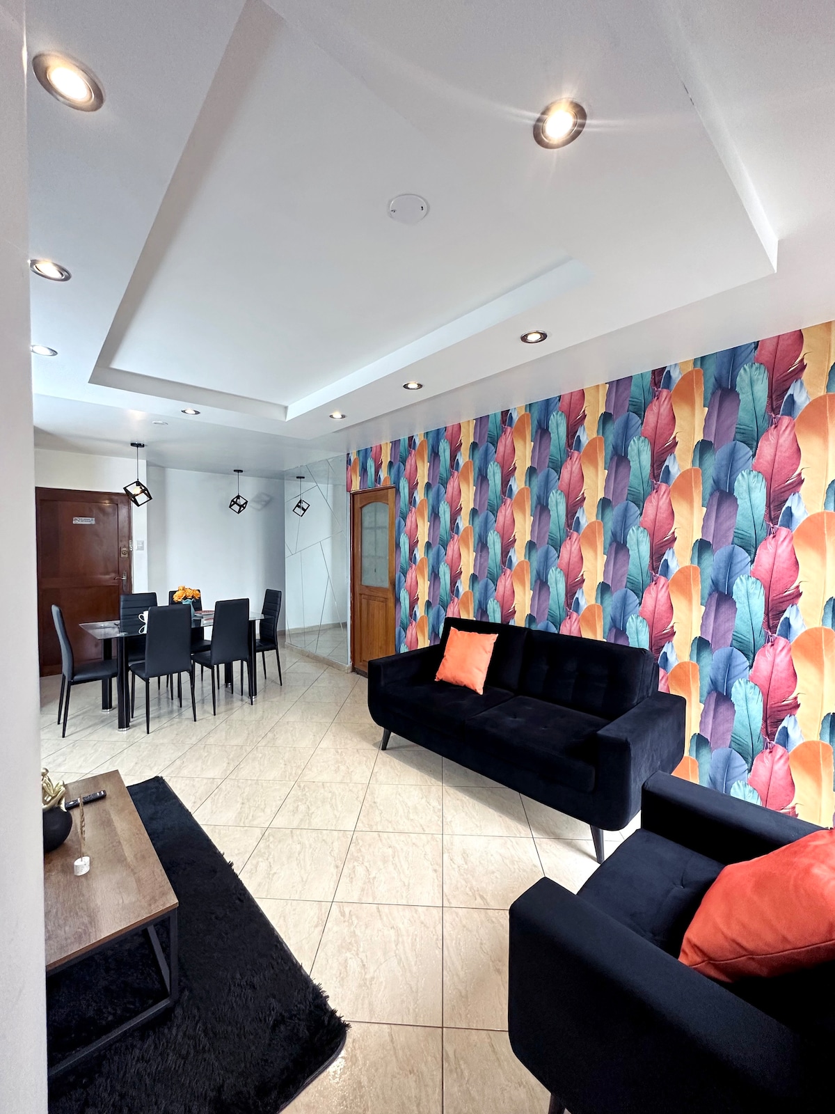 A living area is presented with a stylish black sofa adorned with an orange pillow. A vibrant feather-patterned wallpaper adds color to the space. A wooden coffee table is positioned next to a soft black rug, while a dining area with a dark table and four chairs is visible in the background.
