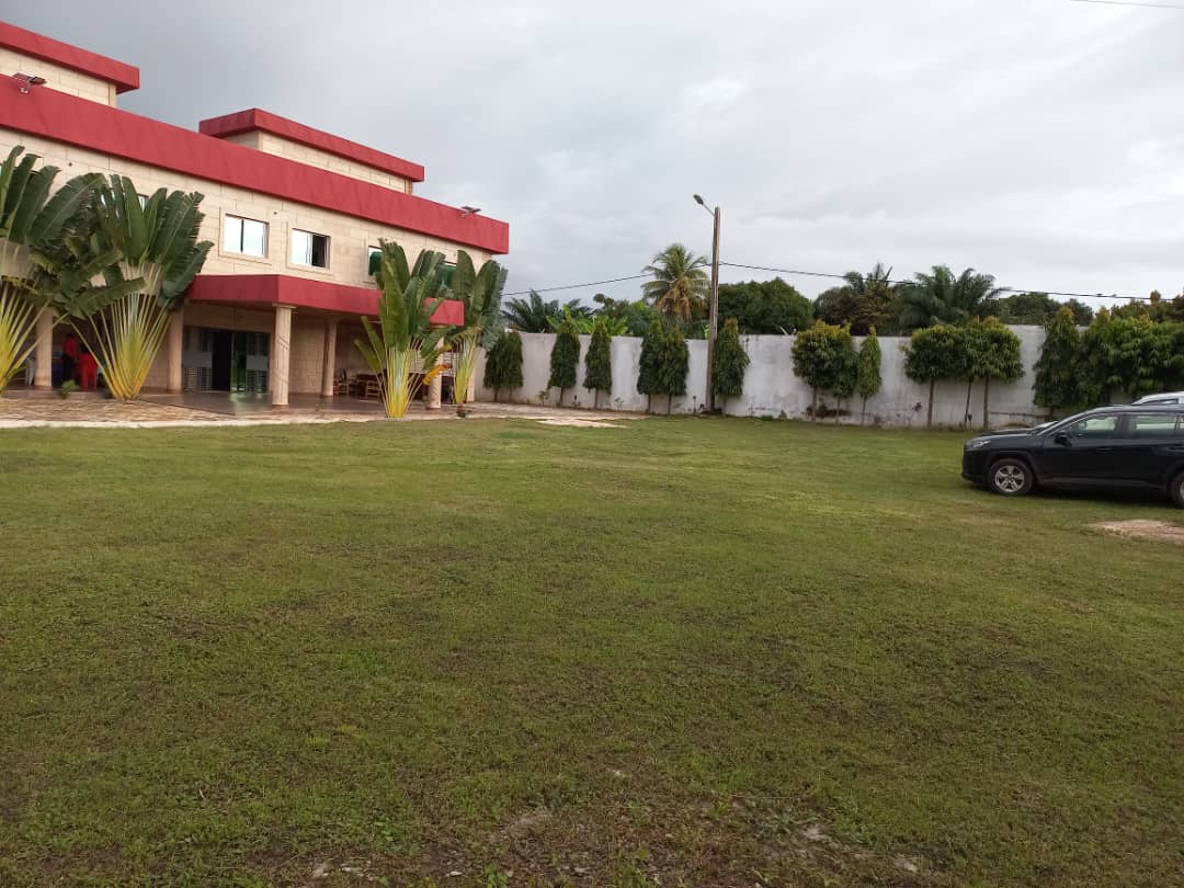 A large grassy area surrounds a two-story building with a red accent roof. Tall tropical plants frame the property, while a dark vehicle is parked on the left side. Lush green trees line the background, complementing the serene outdoor space.