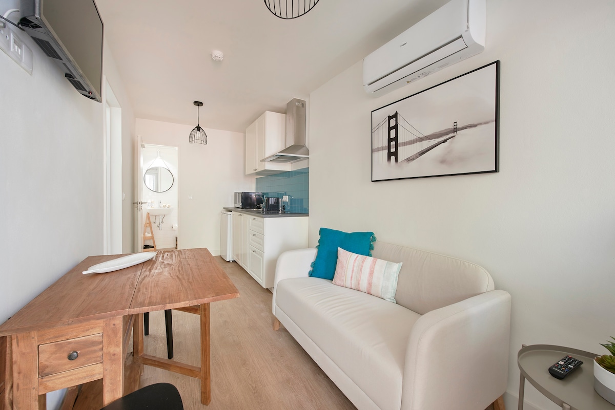 A well-appointed living space features a comfortable sofa with decorative cushions and a wooden dining table. Kitchen appliances are neatly arranged nearby, while a wall-mounted television adds convenience. Natural light brightens the room, complemented by minimalist decor and air conditioning for comfort.