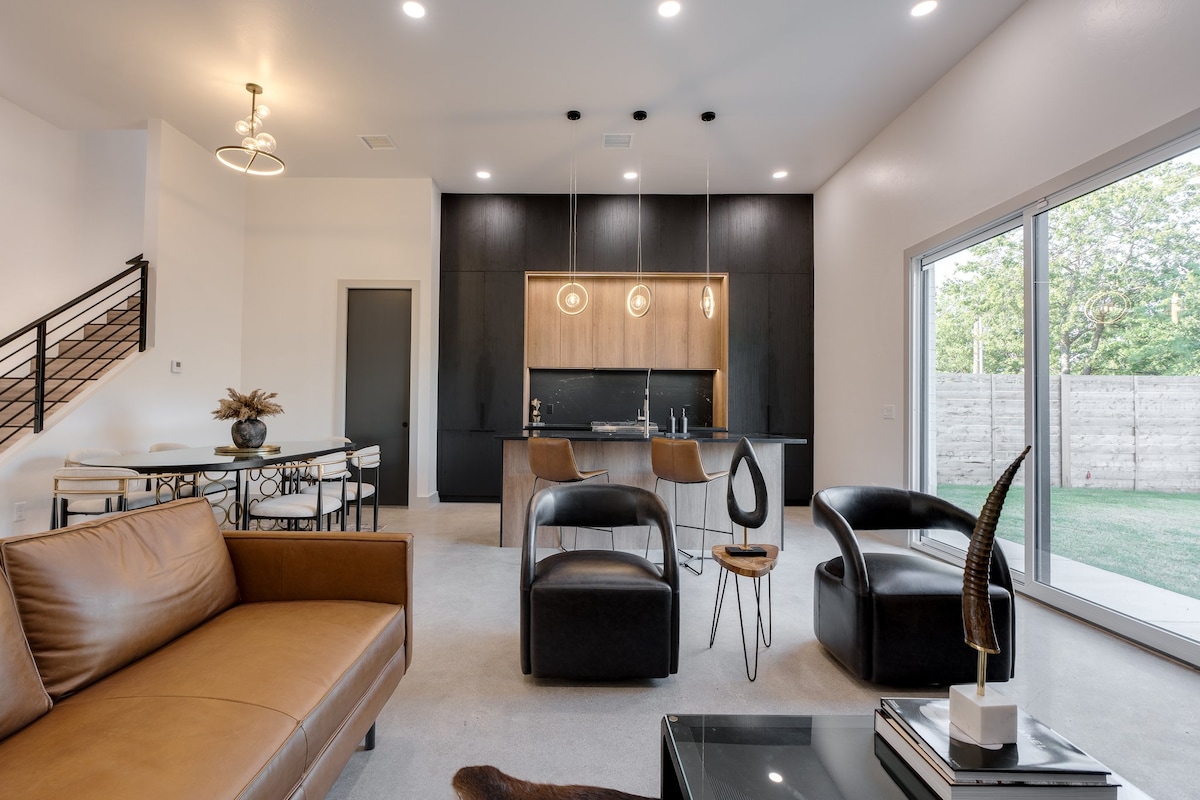 A modern living area is illuminated by overhead lights, featuring a mix of leather and upholstered seating. The stylish kitchen, visible in the background, includes a large island with pendant lighting. A dining table with elegant chairs complements the open layout, and large windows offer a view of the backyard.