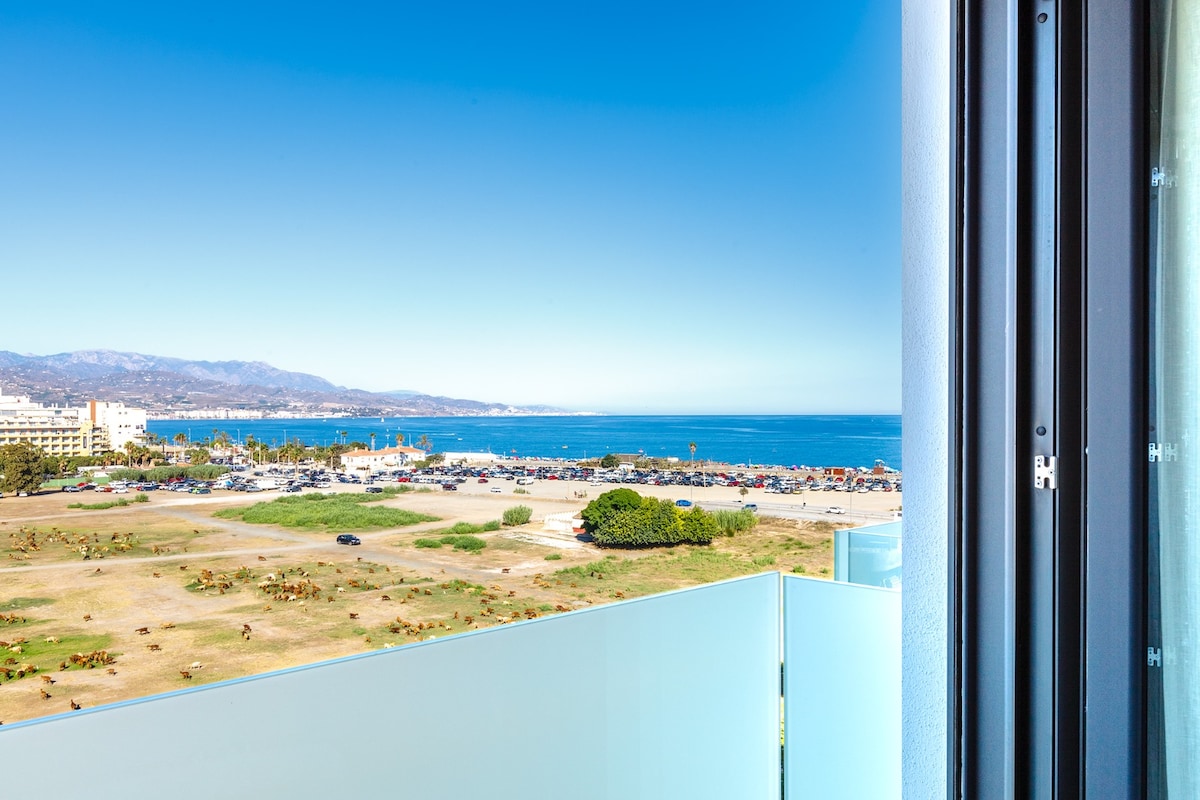A panoramic view of the bay and mountains is visible from the balcony, showcasing clear blue waters and a landscaped area below. The scene captures the vibrant atmosphere of the beach area, with sandy shores and distant coastal activities.