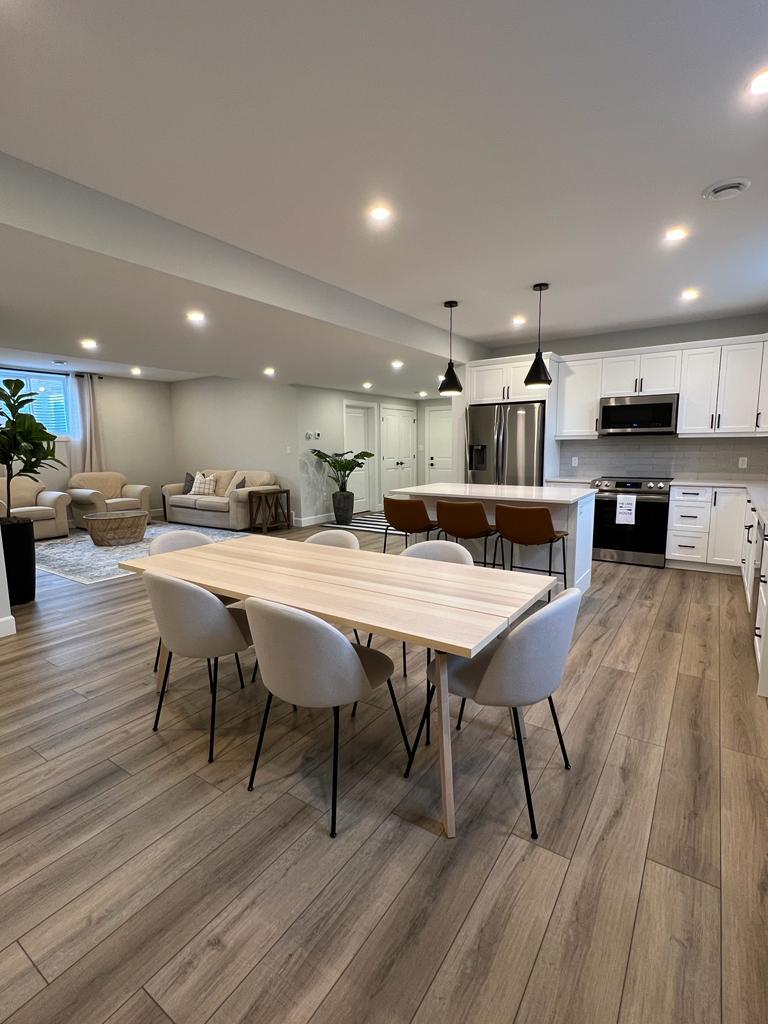 An open-concept living space features a large dining table surrounded by six cushioned chairs. The modern kitchen showcases stainless steel appliances and white cabinetry. A cozy seating area with a light-colored sofa is visible in the background, complemented by decorative plants throughout the room.