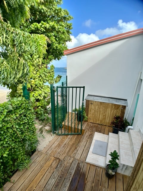A wooden deck leads to a gated entrance, framed by lush greenery. A small storage area is visible beside potted plants. The clear sky hints at the nearby beach, evident just beyond the gate.