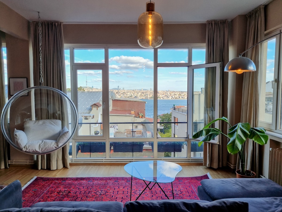 A bright living area features large windows framing a scenic view of the water and city skyline. A cozy hanging chair is positioned near the window, while a sleek coffee table sits on a patterned rug. A plant adds a touch of greenery to the space.