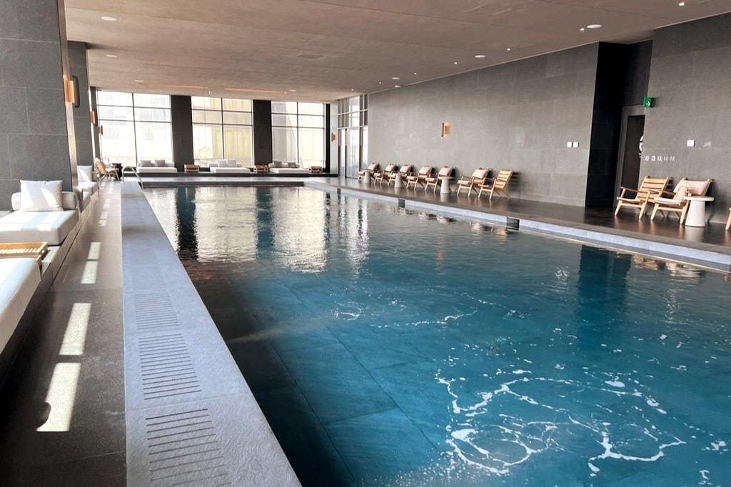 An indoor pool area features sleek gray stone walls and a spacious pool with clear blue water. Sunlight filters through large windows, illuminating the space. Comfortable lounge chairs are arranged along the sides, creating a relaxing atmosphere for guests.