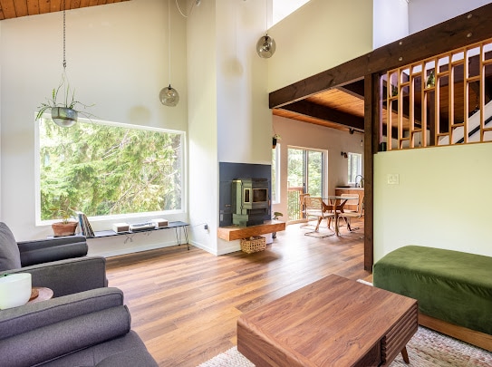 A bright living area showcases a cozy sofa and a wooden coffee table on the warm-toned floor. A modern fireplace is set against a feature wall, with large windows providing views of the surrounding trees. The dining area and loft are visible in the background.