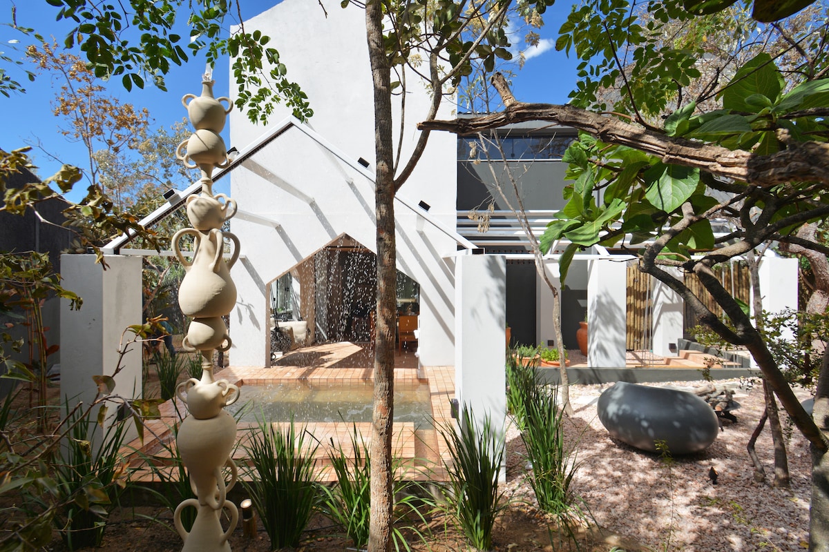 A contemporary structure features an arching entryway lined with lush greenery. A calming water feature is visible, surrounded by decorative stones and plants. Elegant clay sculptures are positioned thoughtfully amidst the garden, enhancing the serene environment.