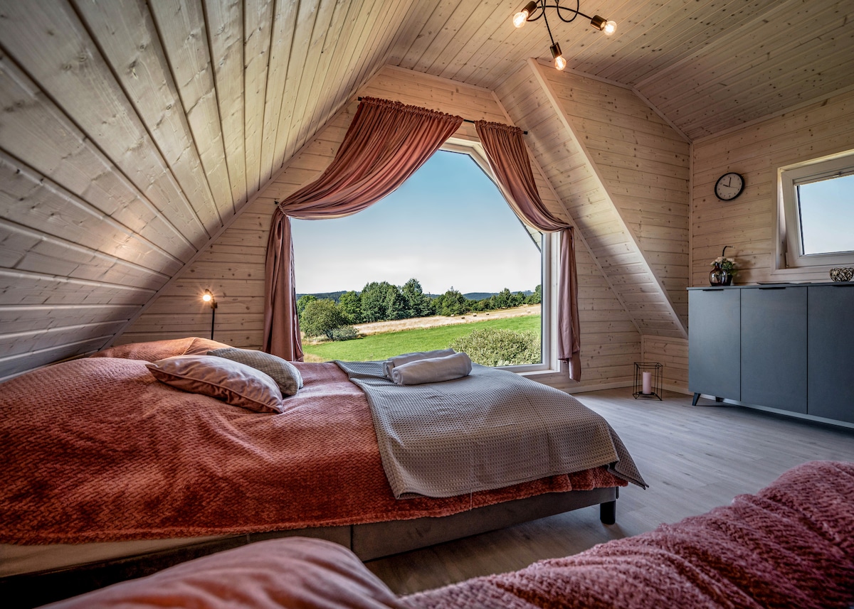 A bright bedroom featuring sloped wooden ceilings and large windows providing expansive views of the landscape. Two spacious beds are dressed in soft, textured linens, with a decorative throw placed on one. Natural light floods the room, enhancing the serene atmosphere.