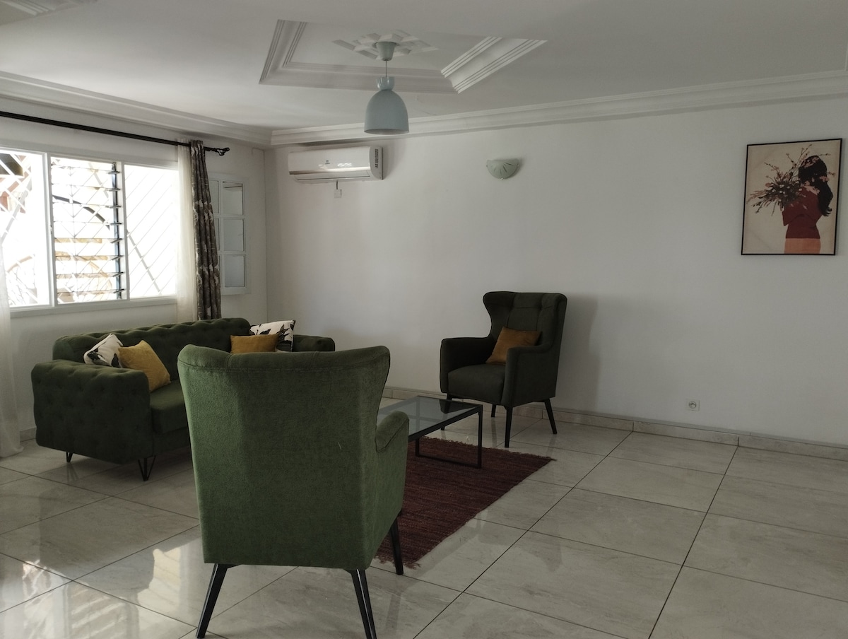 A bright living area is presented with green upholstered armchairs and a small glass coffee table. A neutral-toned rug anchors the seating arrangement on shiny tiled floors. Natural light filters in through a window adorned with light curtains, complementing the minimalist aesthetic.