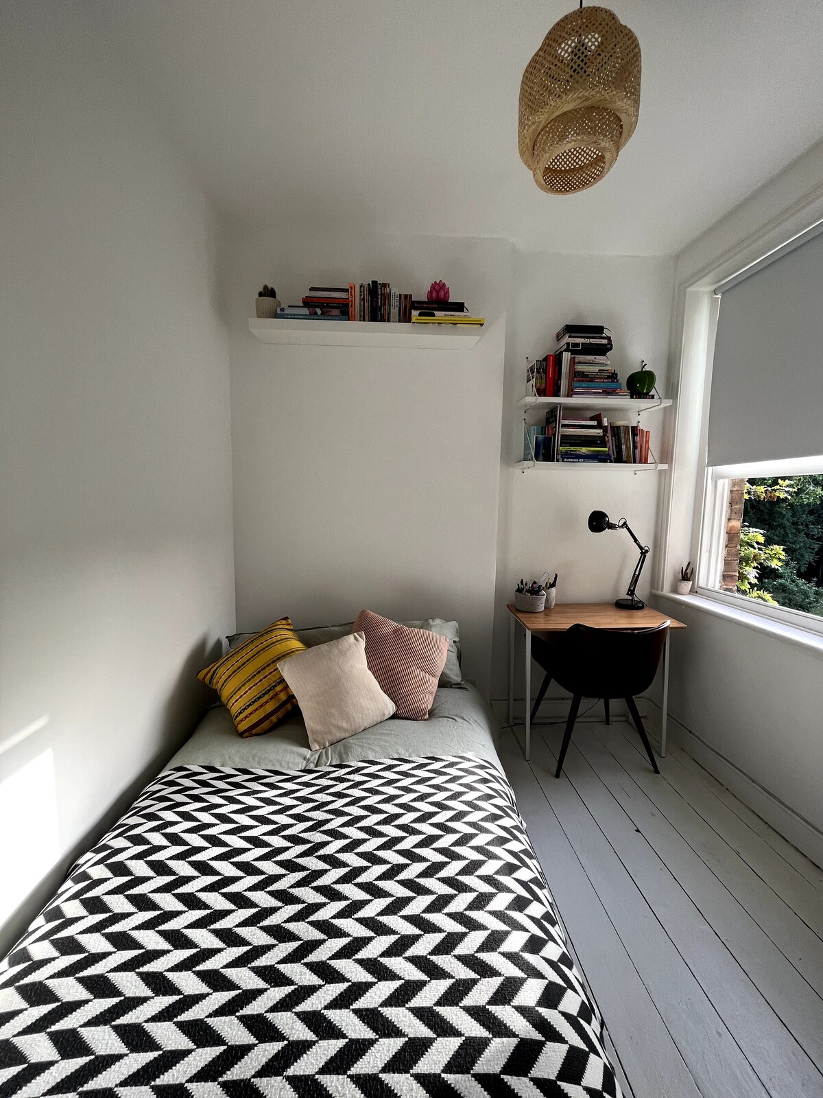 A serene bedroom features a compact single bed dressed in a geometric patterned blanket. A collection of books is displayed on a shelf above. Natural light filters through the window, illuminating a small desk accompanied by a chair, creating a functional workspace.