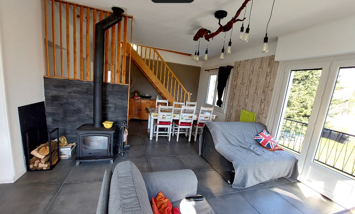 A spacious living area features a wood-burning stove and comfortable seating options. A dining area with several chairs is present, alongside a staircase leading to the upper level. Large windows allow natural light to fill the room, offering views of the outdoors.