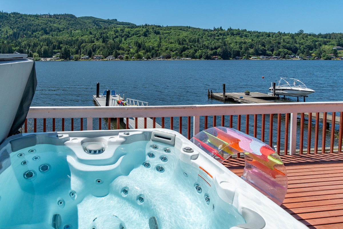 A hot tub is positioned on a sunlit deck, showing clear water and seating for relaxation. In the background, a serene lake reflects the blue sky, with a private dock extending into the water. Lush greenery lines the far shore, enhancing the tranquil lakeside setting.