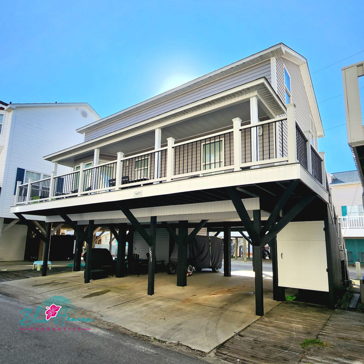 A spacious two-story house is presented with a large front porch supported by sturdy columns. Ample parking space is visible beneath the elevated structure, featuring a concrete driveway. The exterior is finished in light tones, complemented by black railings and trim.