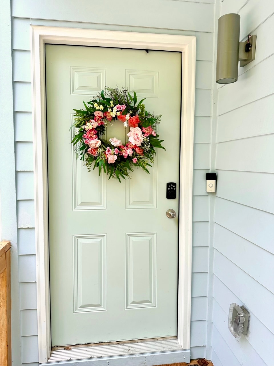 A light green front door is adorned with a floral wreath featuring pink and white flowers. A small keypad for keyless entry and a light fixture are visible on the wall beside the door, which opens to a welcoming atmosphere.