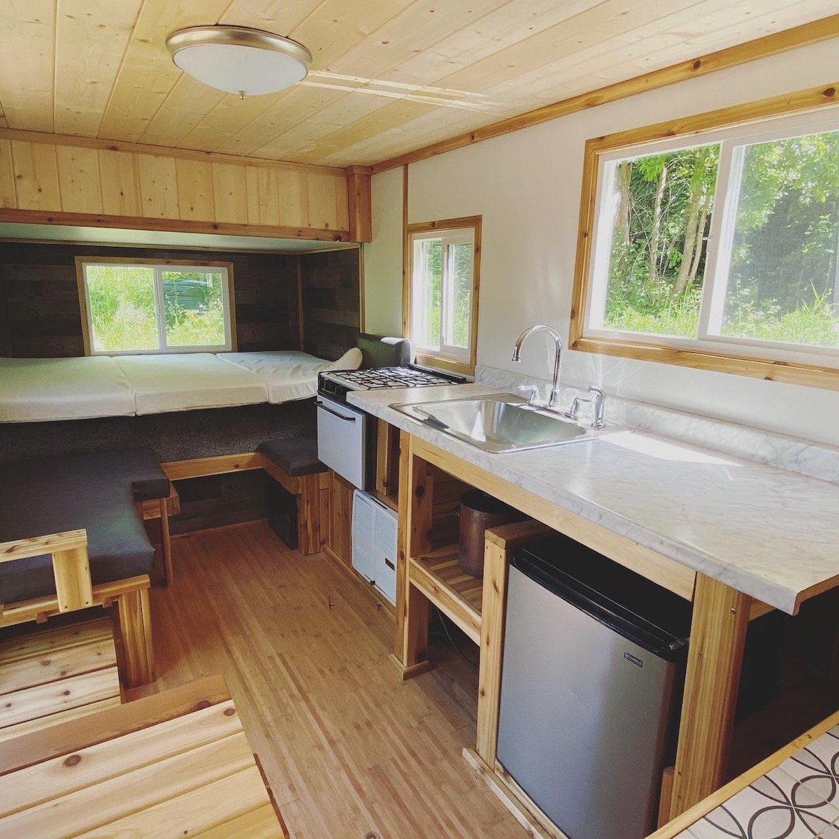 The interior of the tiny trailer showcases a modern kitchen area with a gas stove and oven, a sink with running water, and a mini fridge. Natural light enters through two windows, illuminating the cozy seating and spacious sleeping area, complemented by wooden accents throughout.
