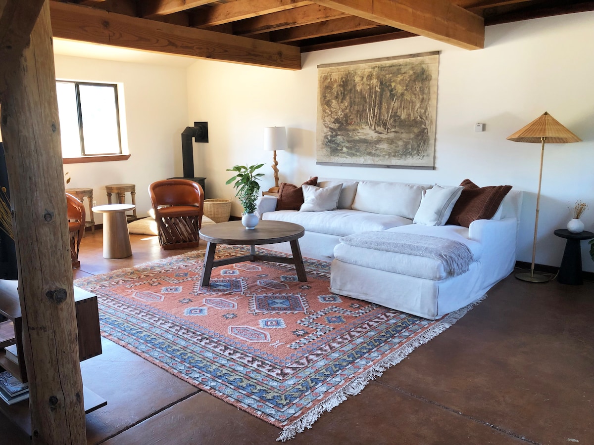 The living area features a comfortable sofa with a chaise, complemented by leather armchairs. A coffee table sits on an area rug, and a floor lamp provides soft lighting. Natural light enters through a window, while rustic beams add character to the ceiling.