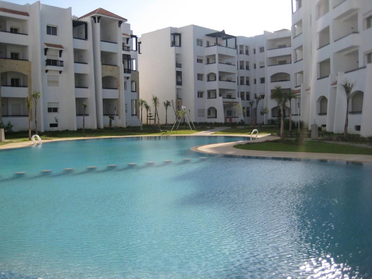 A serene swimming pool is featured in the foreground, surrounded by landscaped green areas. The modern buildings are visible in the background, showcasing light-colored façades and multiple balconies. Palm trees are positioned throughout the landscape, contributing to the overall relaxed atmosphere.