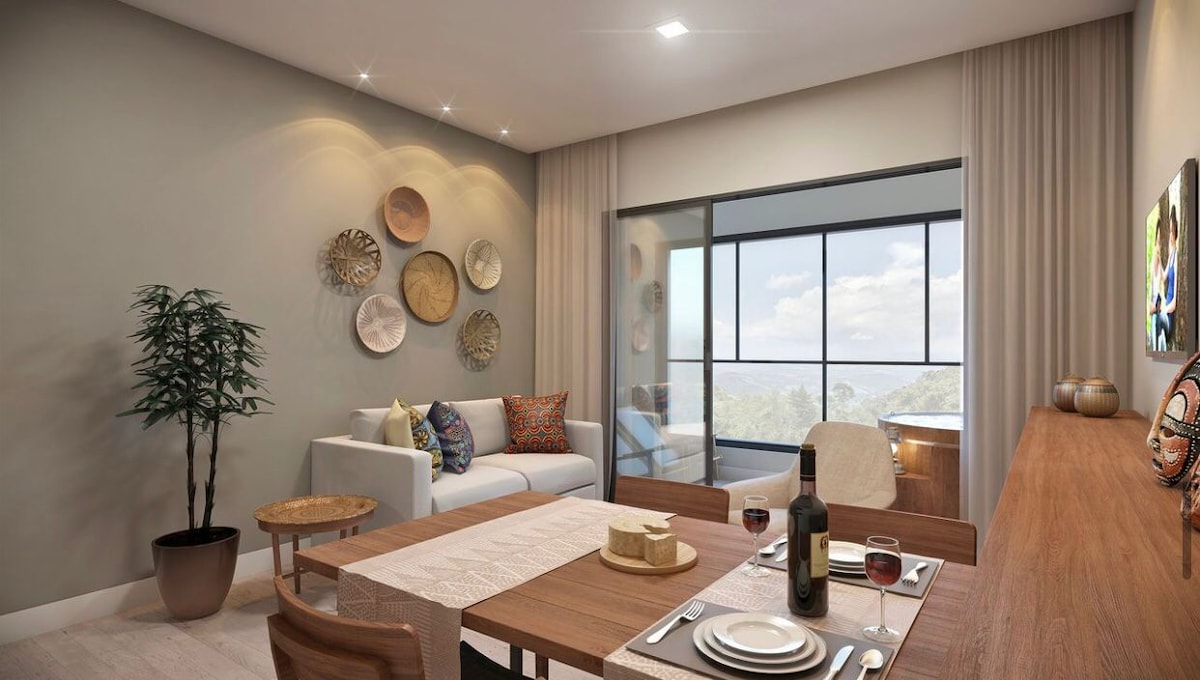 A cozy dining area features a wooden table set for two, complemented by wine glasses and a cheese platter. A comfortable sofa is positioned nearby, adorned with colorful cushions. Decorative plates are arranged on the wall, and large windows provide a view of the outdoors.