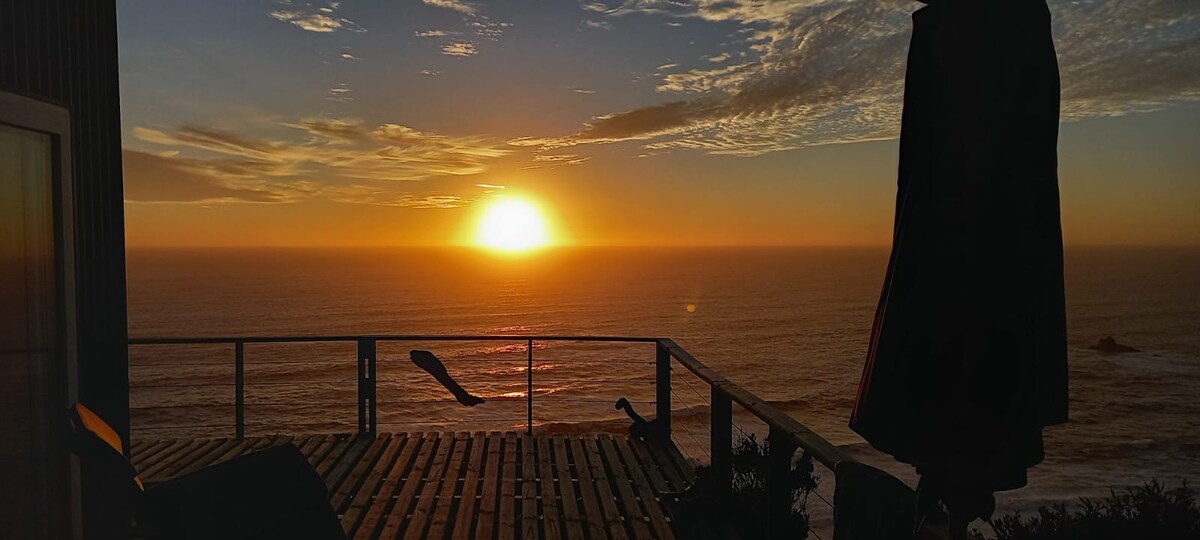 A breathtaking sunset is captured over the ocean, casting warm hues across the sky and water. Silhouettes of a railing and outdoor furniture frame the scene, highlighting the serene surroundings of the coastal setting.