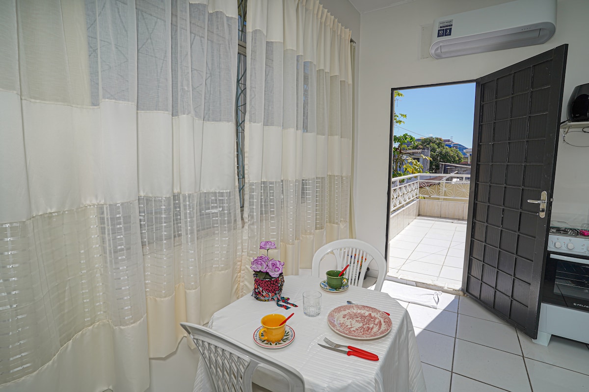 A bright dining area is visible, featuring a small table set for two. White curtains gently filter the light, while an open door leads to a private balcony. The kitchen area shows a glimpse of the stove near the entrance.