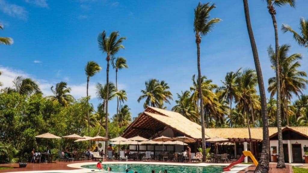 The outdoor area features a swimming pool surrounded by lush palm trees and lounge chairs. A large thatched-roof structure serves as a social space offering shade. Families enjoy recreational activities around the pool, highlighting a vibrant atmosphere with sunny skies.