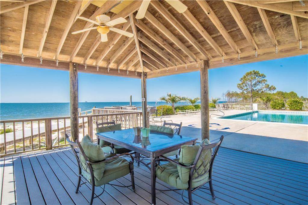 An open-air pavilion features a spacious dining table surrounded by comfortable seating. A ceiling fan offers a gentle breeze, while the views of the beach and pool extend beyond. Natural wood beams highlight the structure, offering a welcoming space for gatherings.
