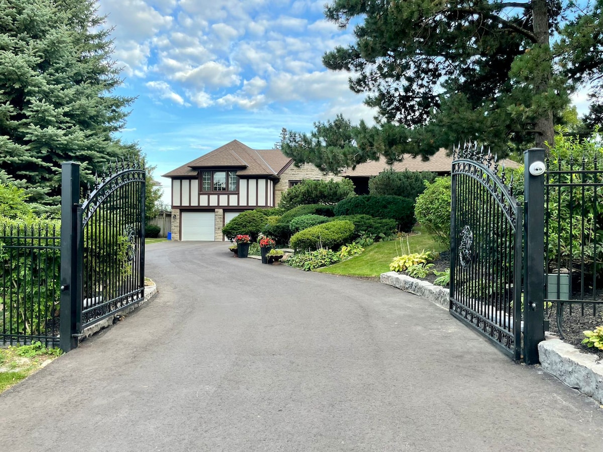 A grand entrance is highlighted by ornate wrought iron gates opening to a well-maintained driveway. Lush greenery and colorful flower beds frame the path, leading to a spacious two-story home with a sloped roof and inviting exterior. A clear sky provides a bright backdrop.
