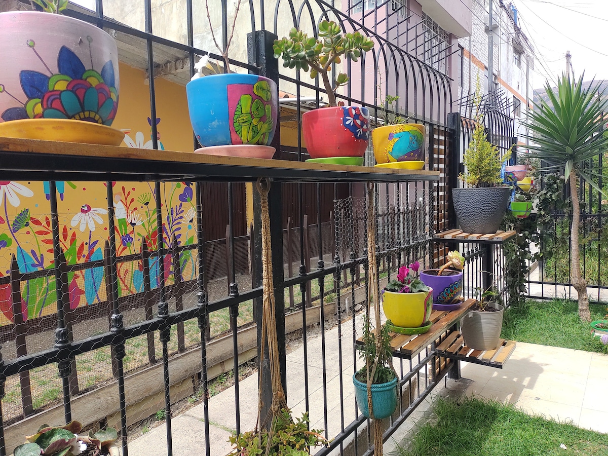 Colorful potted plants are arranged along a wooden shelf against a metal fence. Various pots in vibrant hues are decorated with artistic patterns, complementing a mural of flowers on the wall behind. The area is enhanced by a lush green lawn.