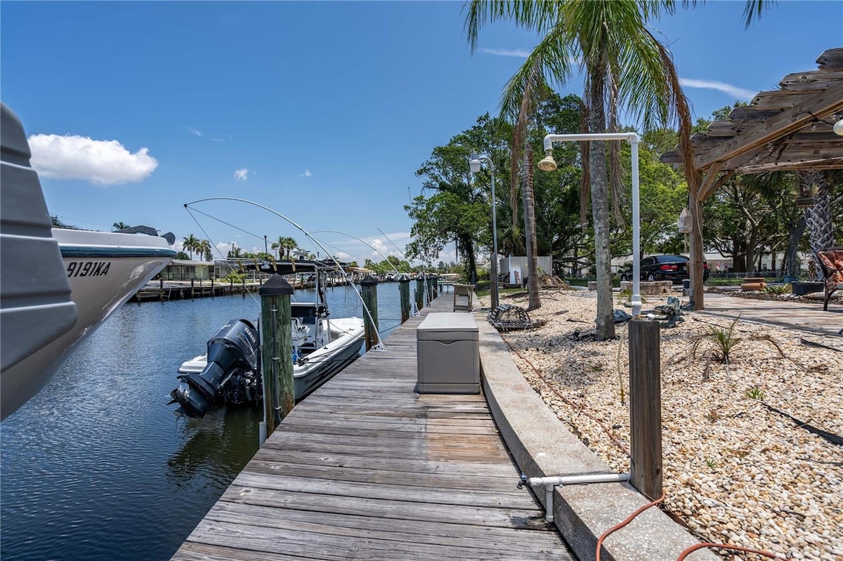 The wooden dock extends alongside the calm canal, flanked by palm trees and sandy landscaping. A boat is moored nearby, and the area features light-colored gravel and outdoor fixtures. The scene is illuminated by bright sunlight, enhancing the tranquil setting.