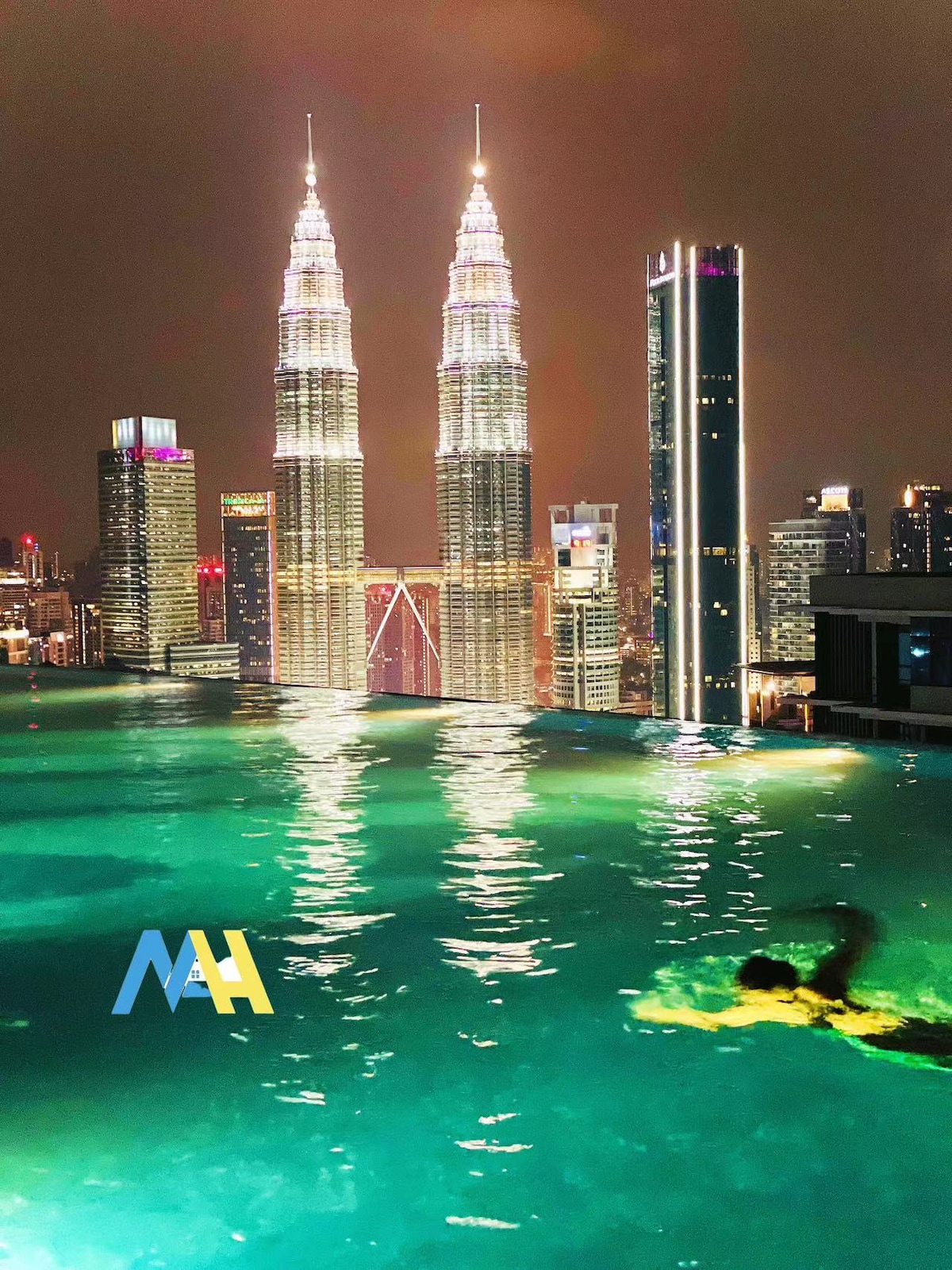 An infinity pool is depicted at night, creating a seamless view of the city skyline featuring the illuminated Petronas Twin Towers. Gentle ripples in the water reflect the surrounding lights, enhancing the tranquil atmosphere. A swimmer is seen gliding through the pool.