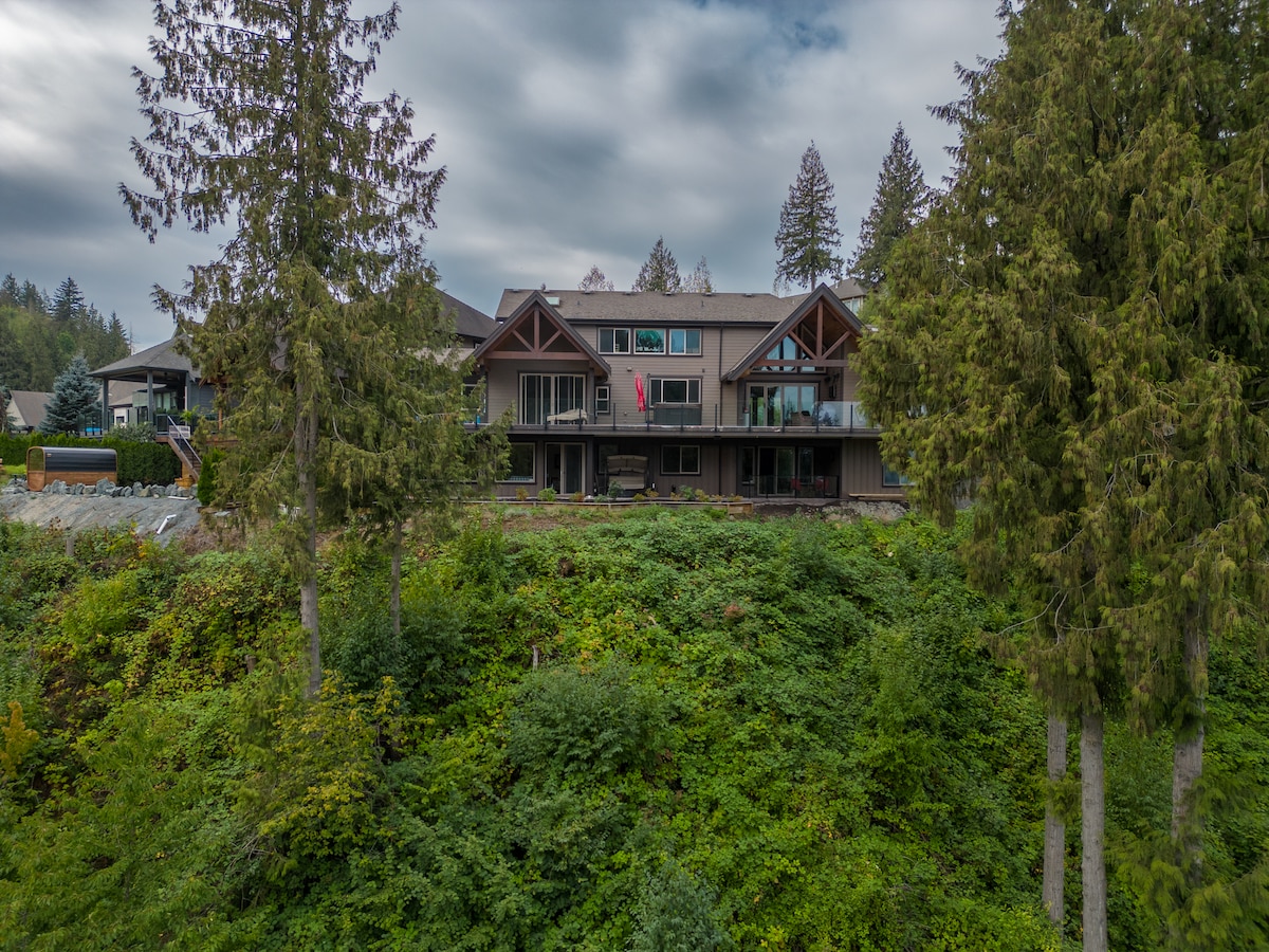 The exterior of a multi-level house is nestled among tall trees, with a backdrop of lush greenery. The architecture features a mix of wood and stone elements. Several balconies are visible, showcasing a welcoming atmosphere. A flag hangs from one of the balconies.