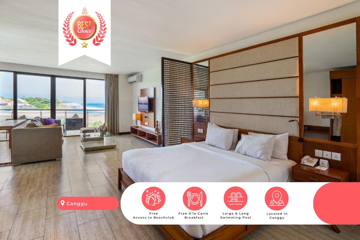A spacious suite is presented, featuring a comfortable double bed with crisp white linens. A well-lit living area with modern furnishings is visible, along with large windows offering views of the Indian Ocean. A flat-screen television is positioned on a wooden unit.