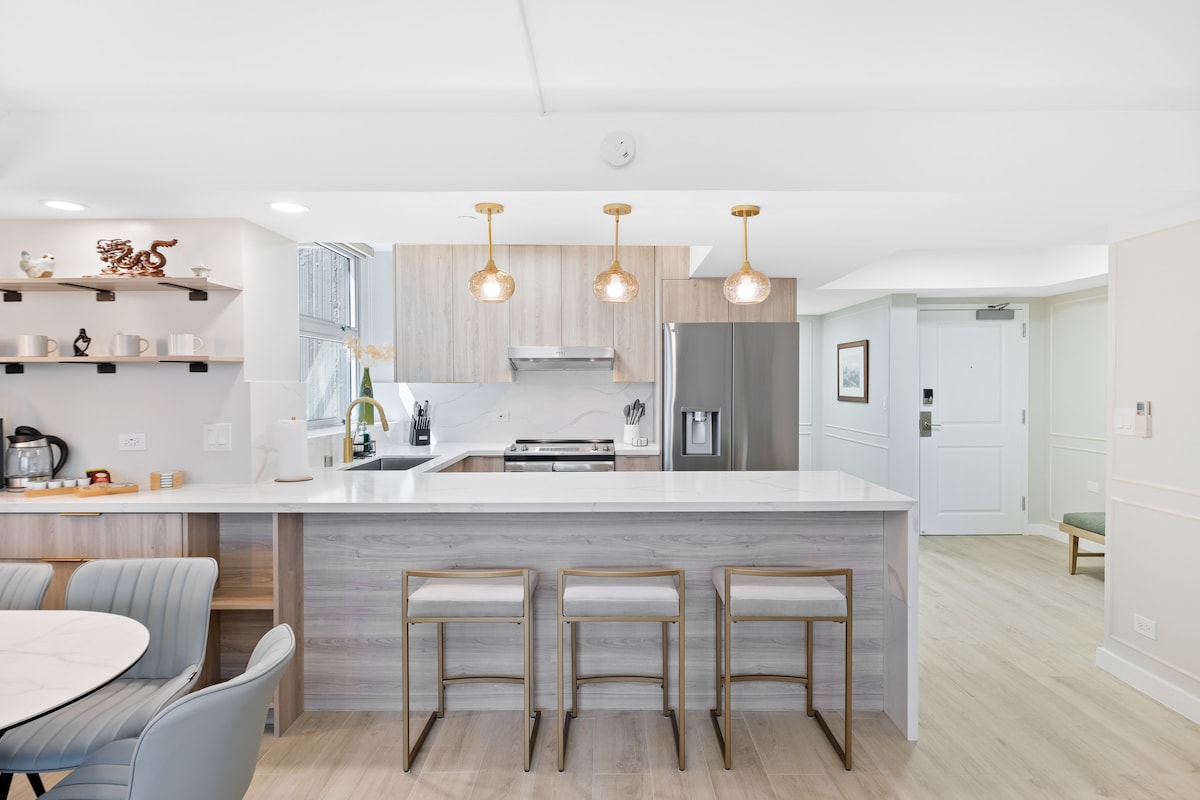 A sleek kitchen with modern finishes is showcased, featuring a large white countertop and stainless steel appliances. Three bar stools are positioned at the counter, illuminated by pendant lighting. A dining area with light grey chairs complements the bright, open space.