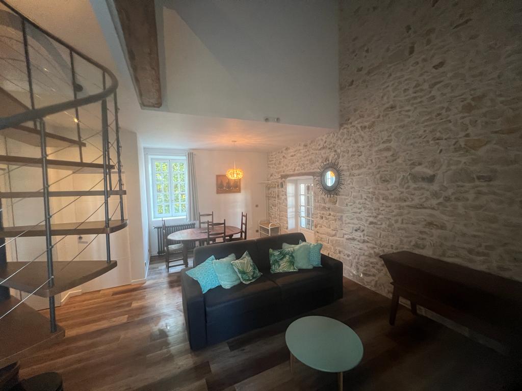A living area features exposed stone walls and warm wooden flooring. A spiral staircase leads to an upper level. A black sofa adorned with decorative pillows sits near a round table, while natural light enters through the windows, enhancing the space.