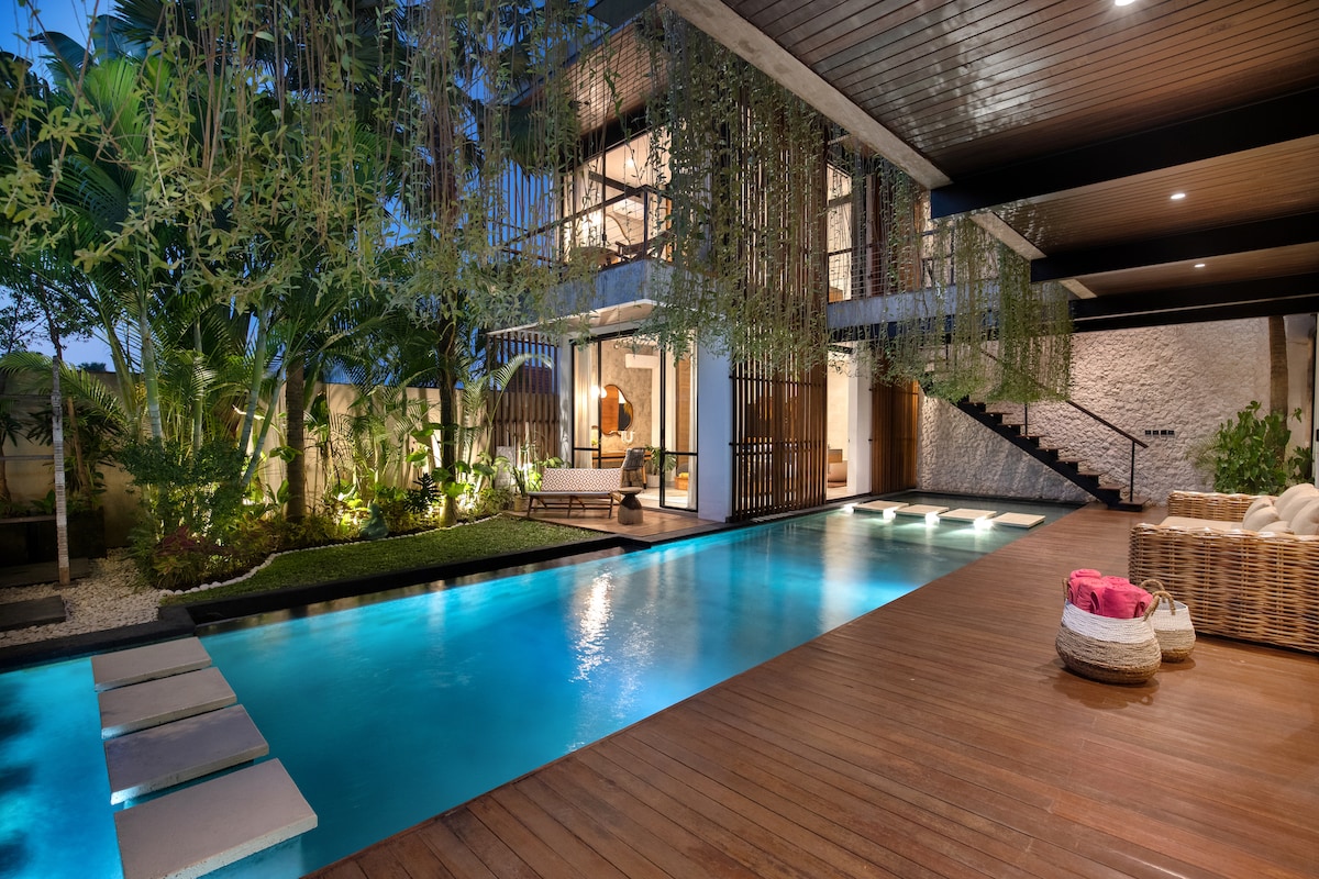 A modern villa showcases an inviting private pool framed by wooden decking and lush greenery. The tranquil water reflects the surrounding architecture, with an open living space visible in the background. Soft lighting enhances the serene atmosphere in the early evening.