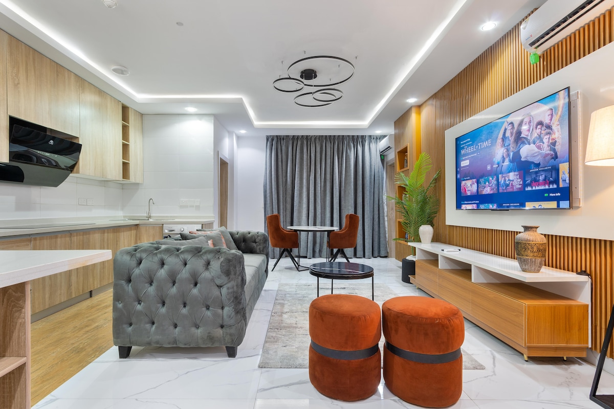 The living area is well-lit, featuring a modern design with a tufted grey sofa and two round ottomans. A large television is mounted on the wall, displaying a colorful interface. The space includes a dining table set for four, with plants adding a touch of greenery.