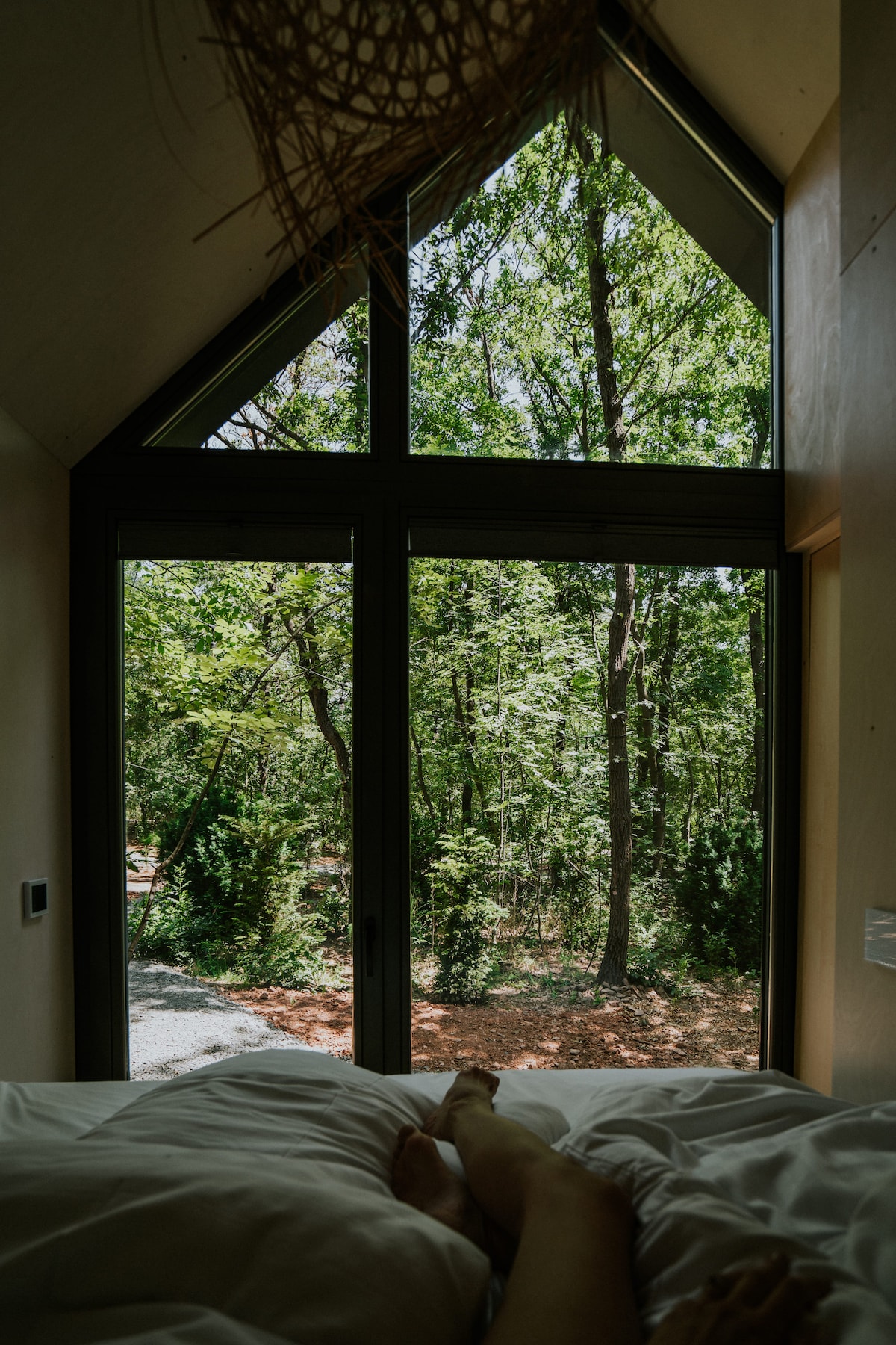 A spacious bed is positioned near large triangular windows, offering a view of the surrounding trees and forest. Soft, natural light filters in, creating a serene atmosphere, while a cozy bedding arrangement invites relaxation.