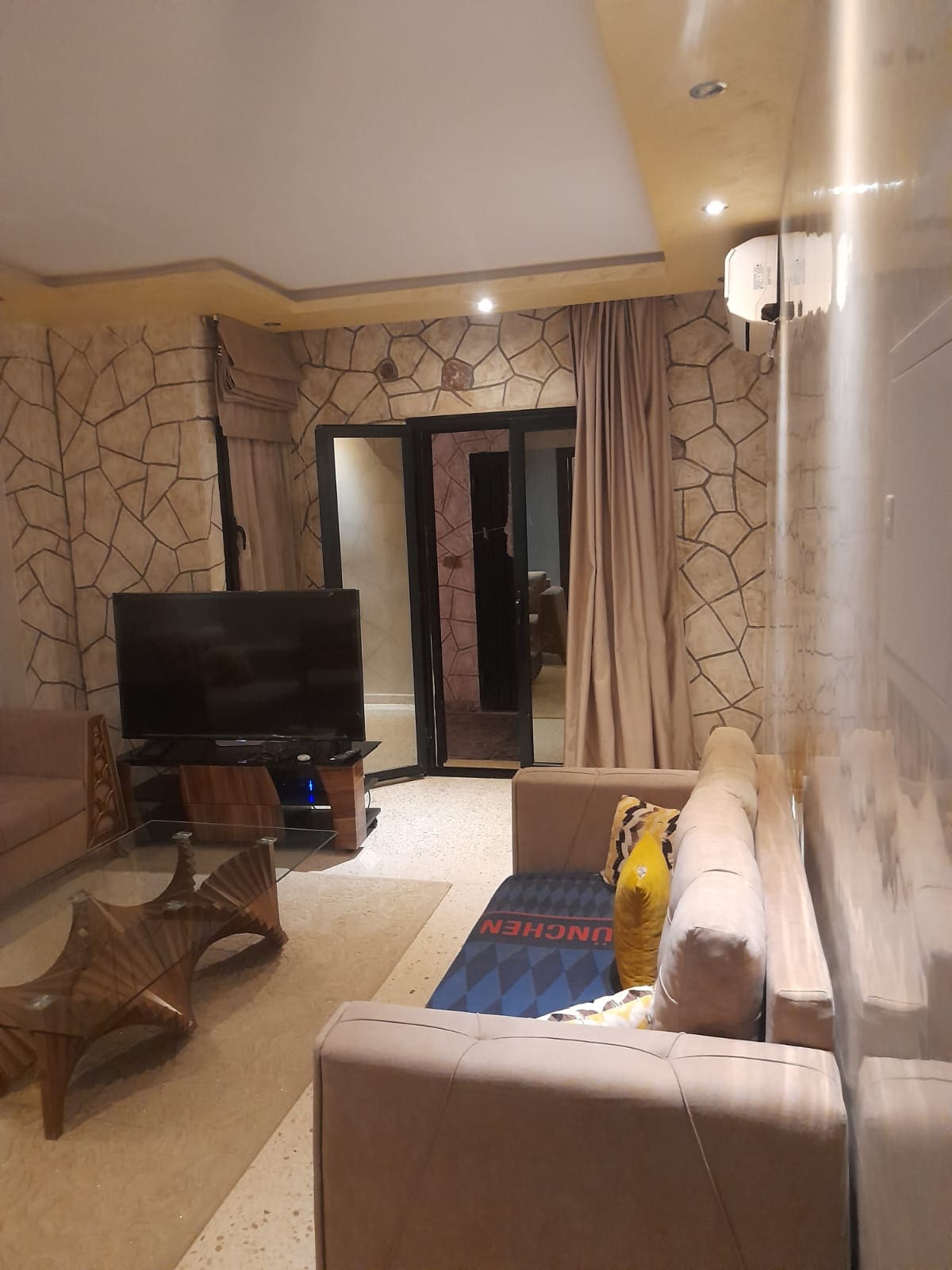 A relaxed seating area features a plush sofa and a glass coffee table. A large television is positioned against a textured wall. Light-colored curtains frame the glass doors, leading to an outdoor space. Soft lighting enhances the warm tones of the room.