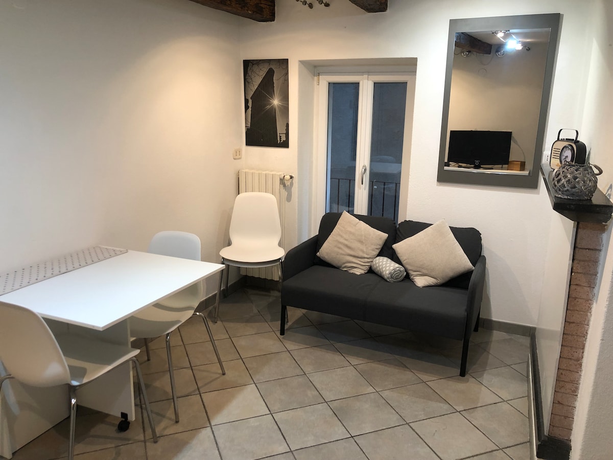 A compact living area is displayed, featuring a dark gray sofa with two decorative cushions. A dining table with four white chairs is positioned nearby. A mirror and framed artwork adorn the wall, while a window offers natural light, contributing to the clean, neutral color scheme.